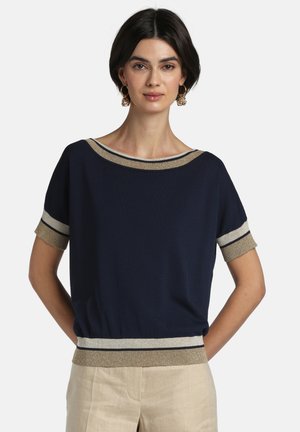 Kvinde iført en marineblå kortærmet top med guld- og hvide kanter ved krave, ærmer og kant, kombineret med lyse beige bukser.