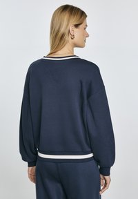 Felpa navy con una vestibilità rilassata, caratterizzata da colletto e polsini a costine, strisce bianche decorative sull'orlo e una superficie in tessuto testurizzato.