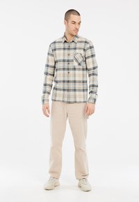 Knoopdoorgeknoopte geblokte blouse in beige, blauw en bruin; voorzien van een borstzak en lange mouwen. Gecombineerd met lichtbeige broek en sneakers.