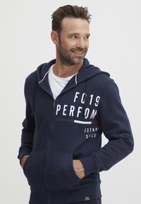 Sudadera con cremallera en color navy, con un bolsillo tipo canguro, estampado de logo en blanco y detalles a rayas rojas y blancas en la capucha. Material suave de mezcla de algodón.