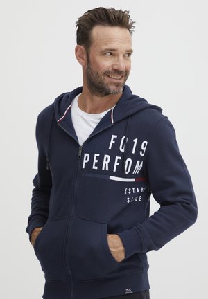 Navy zip-up hoodie with a kangaroo pocket, white logo print, and red and white striped accents on the hood. Soft cotton blend material.
