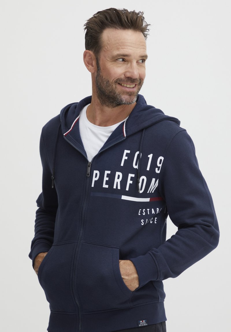 Sudadera con cremallera en color navy, con un bolsillo tipo canguro, estampado de logo en blanco y detalles a rayas rojas y blancas en la capucha. Material suave de mezcla de algodón.