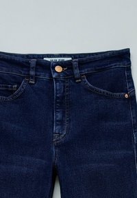 Jeans de mezclilla azul oscuro con un diseño ajustado, que cuentan con un cierre de botón de cobre y costuras amarillas. La etiqueta dice "Salsa Jeans."
