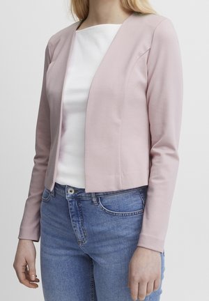 Vrouw die een lichtroze blazer met open voorkant draagt, een wit geribd topje en een blauwe spijkerbroek, staand tegen een simpele lichte achtergrond.