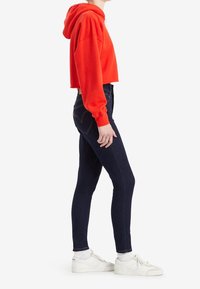 Sudadera roja recortada con cordón, combinada con vaqueros ajustados de color azul oscuro que tienen bolsillos traseros y detalles de costura; complementada con zapatillas blancas.