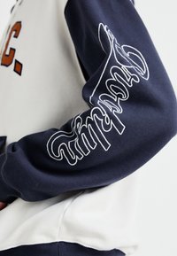 Manche de sweat-shirt bleu marine avec un texte en cursive blanche contourée sur le bras, portée sur un haut crème avec des lettres marron partiellement visibles.