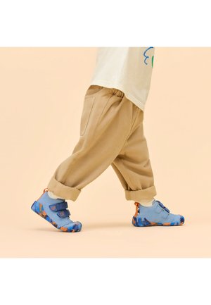 Bambino che cammina indossando pantaloni beige risvoltati e scarpe da ginnastica blu con suole decorate da un motivo arancione e blu su uno sfondo beige.
