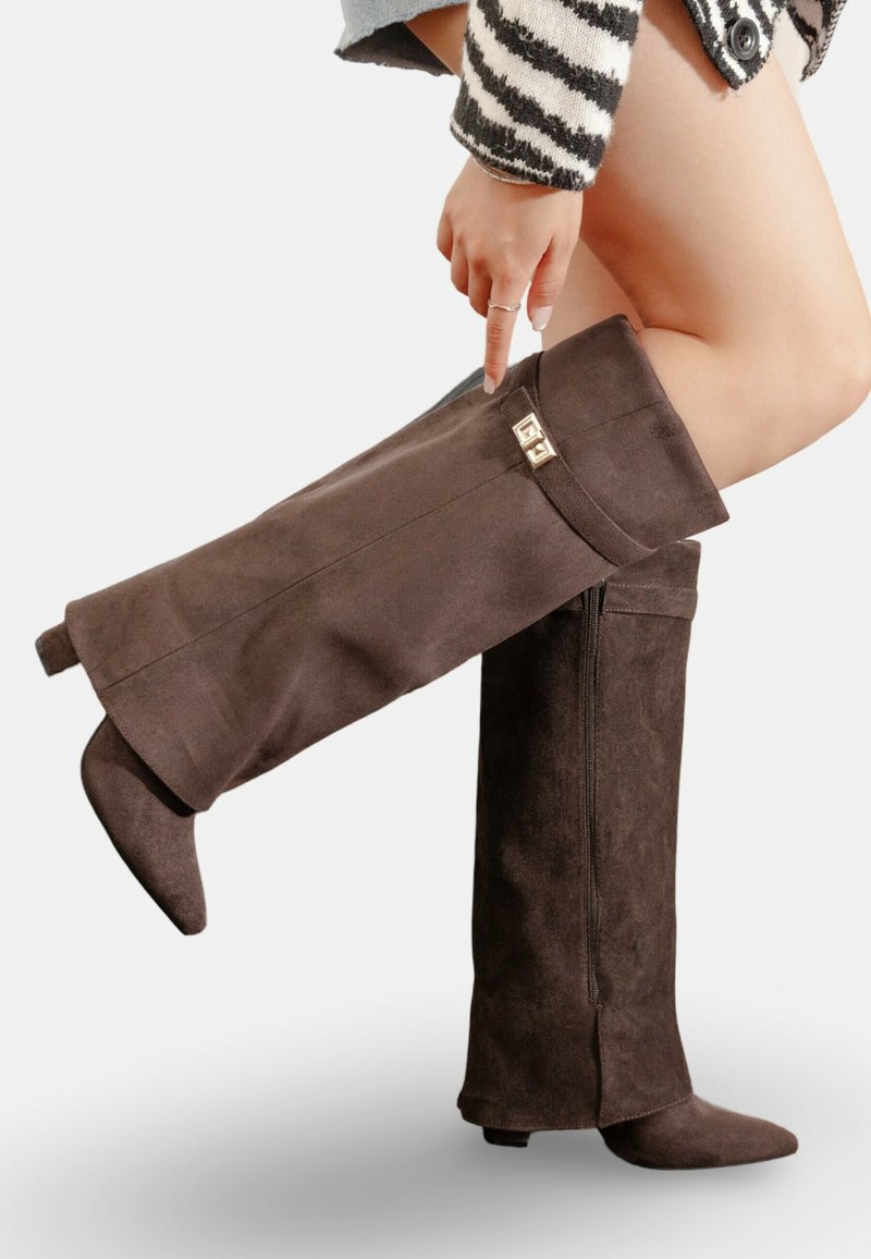 Bottes en suède marron au-dessus du genou avec un bout pointu, un haut replié et des accents métalliques décoratifs sur le revers. Fermeture éclair sur le côté pour plus de facilité à enfiler.