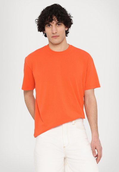 T-shirt arancione brillante in cotone, con collo rotondo e maniche corte. Il tessuto appare liscio; indossato con pantaloni in denim bianchi.
