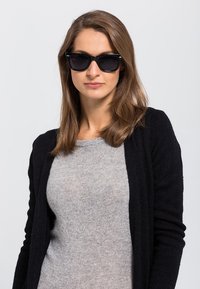 Ochelari de soare negri cu rame largi purtați de o femeie, asociați cu un pulover gri deschis și un cardigan negru, având o textură fină.