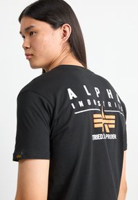 Crna majica kratkih rukava s natpisom "ALPHA INDUSTRIES" u bijelom i narančastim grafičkim logom na leđima. Mekani pamučni materijal, opušteni kroj.