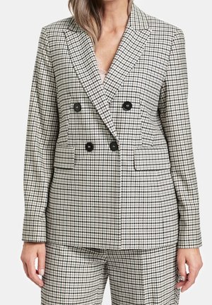 Kobieta ubrana w dwurzędową marynarkę w beżowo-czarną pepitkę z dopasowanymi spodniami, stojąca na neutralnym tle.