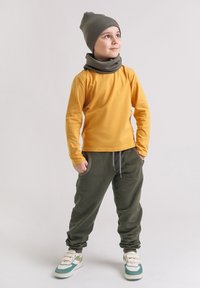 Żółta koszula z długim rękawem, oliwkowozielone spodnie dresowe z kieszeniami, szara czapka typu beanie oraz dopasowany komin. Białe i turkusowe sneakersy z akcentami.