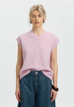 Jeune femme aux cheveux blonds portant un pull sans manches violet clair et un jean bleu taille haute, debout devant un fond uni.