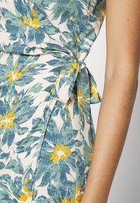 Kremi värviline lilleline mähkimiskleit, millel on sinine ja kollane botaaniline trükk, sidumisse detail külgedel ja tekstureeritud kangas.