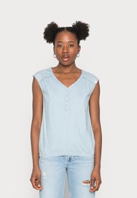 Lys blå denim topp med V-hals, subtil knappelukking og korte cap-ermer. Har delikate sømmer langs halslinjen.