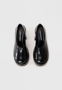 Ζευγάρι μαύρα δερμάτινα παπούτσια τύπου slip-on με χονδρά πέλματα, σε θέα από πάνω σε ανοιχτό γκρι φόντο.