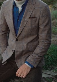 Blazer a cuadros marrón con acentos en azul claro y azul oscuro, que presenta solapa muesca, cierre de botón y pañuelo de bolsillo, superpuesto sobre una camisa de mezclilla.