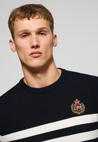 Marineblauer Strickpullover mit weißen horizontalen Streifen, geripptem Rundhalsausschnitt und gesticktem Logo auf der Brust. Weiche Textur, figurbetontes Design.