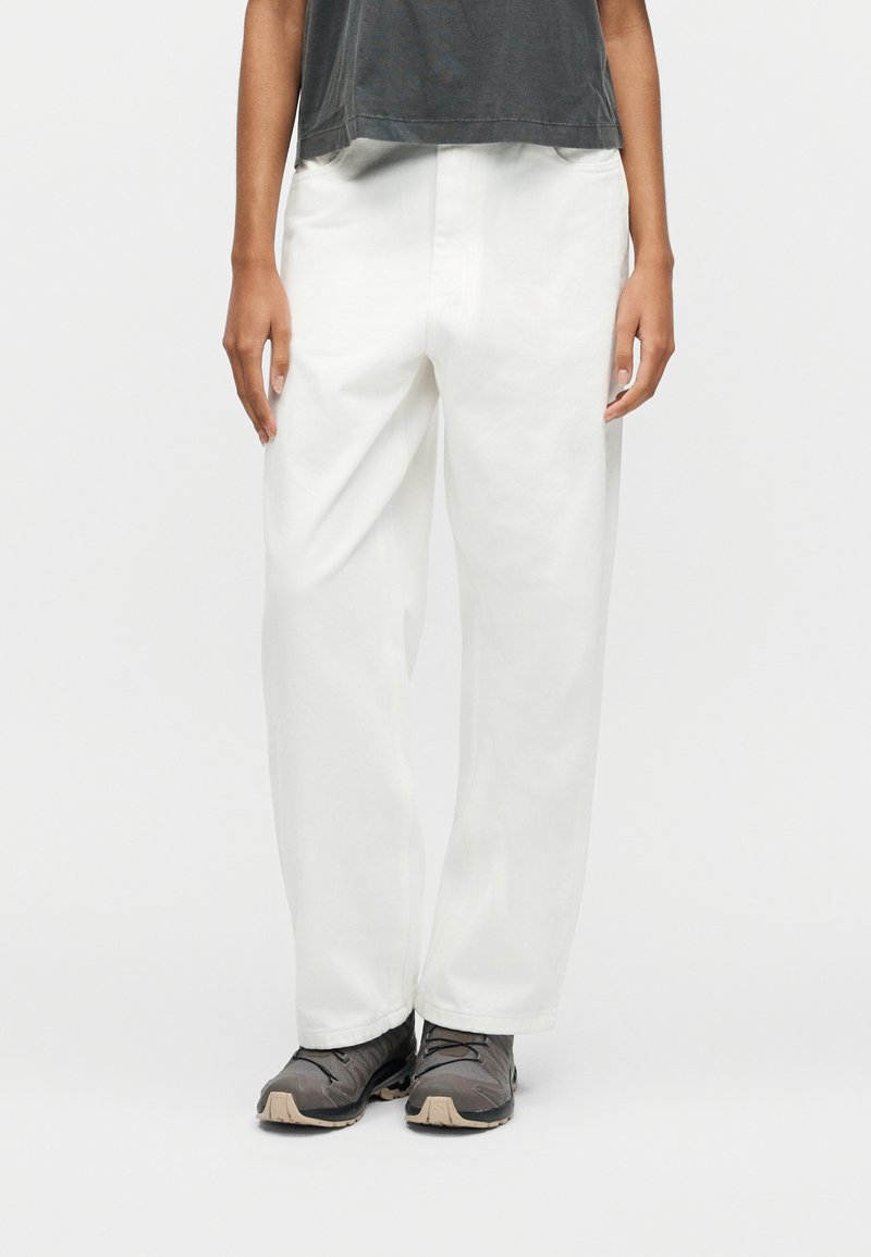 Pantaloni bianchi, ampi, dalla trama liscia. Presentano un design classico, con vita alta, abbinati a scarpe da corsa scure.