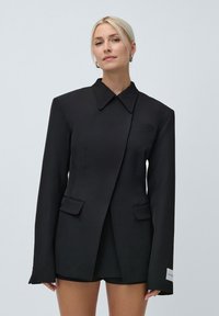 Femeie blondă îmbrăcată într-un blazer negru supradimensionat cu umeri căptușiți și pantaloni scurți negri asortați, stând în fața unui fundal simplu.