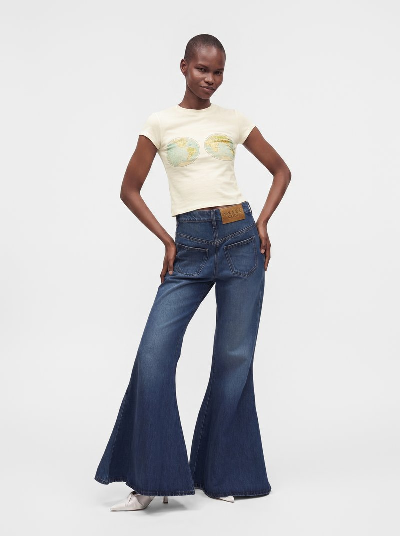 Femme portant un t-shirt crème avec un imprimé carte du monde et un jean bleu taille haute à jambes larges, posant avec les mains sur les hanches devant un fond blanc.