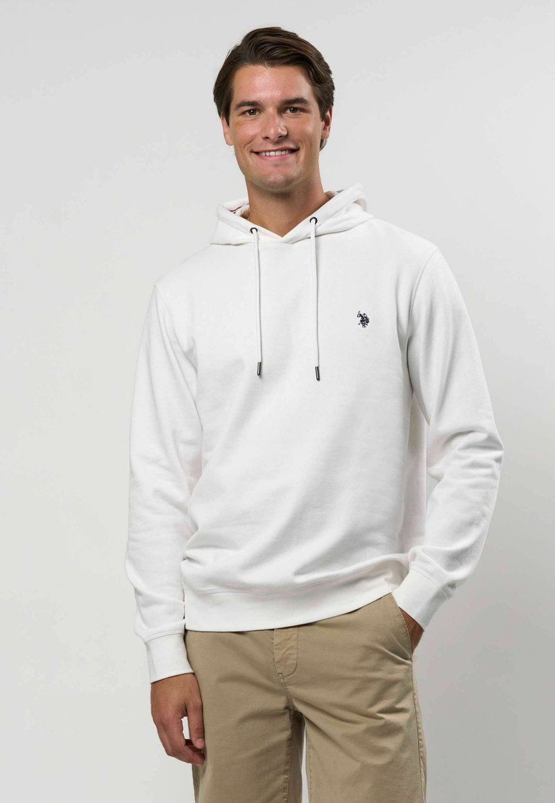 Hoodie blanc avec cordons, poche avant, et petit logo bleu marine sur la poitrine. Tissu lisse, coupe décontractée, porté avec un pantalon beige.