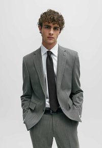 Costume léger gris à rayures avec une veste à un seul bouton et un pantalon assorti. Chemise blanche, cravate noire texturée et ceinture noire.
