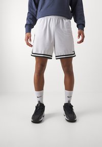 Calções desportivos brancos com riscas pretas, cintura elástica e textura de malha, combinados com sapatilhas pretas e meias brancas com um logótipo.