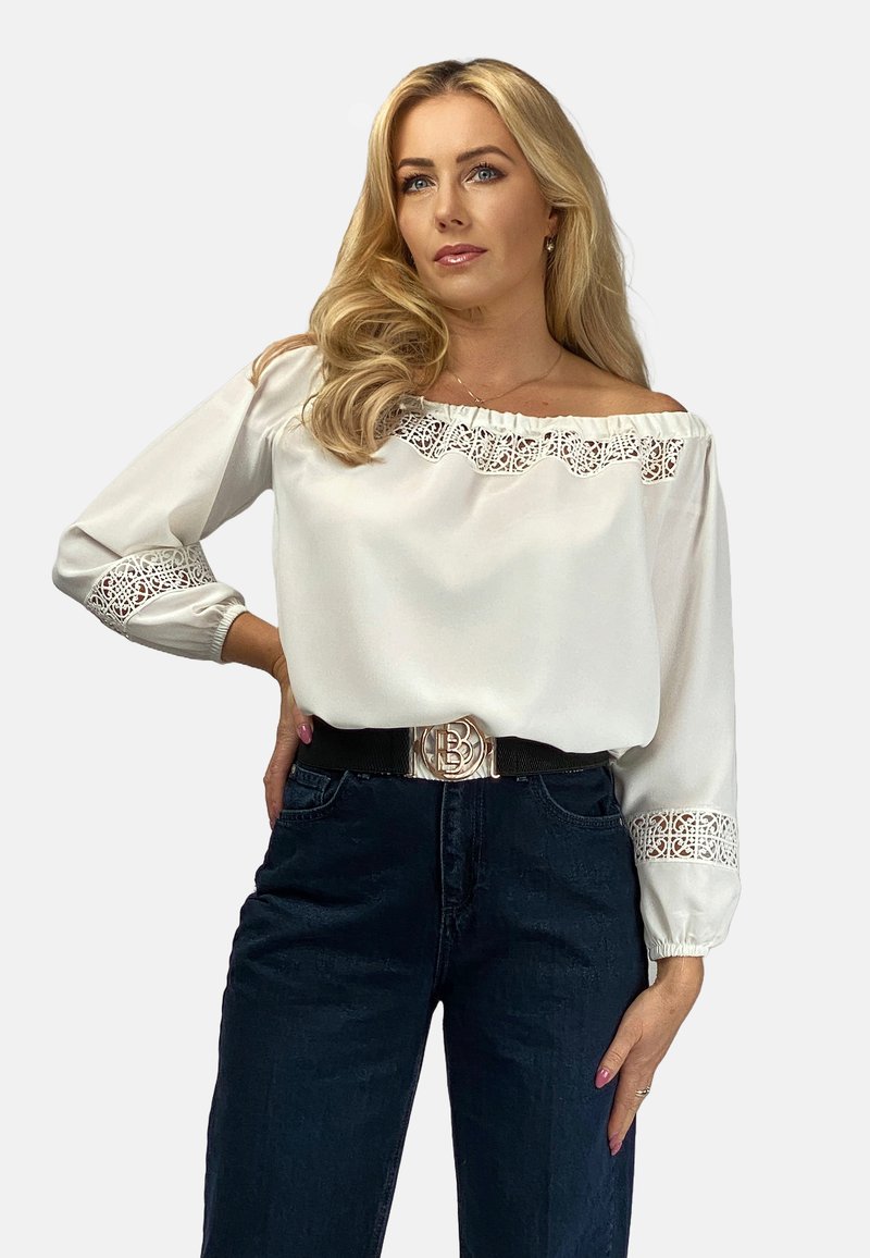Biała bluzka z dekoltem off-shoulder, z koronkowymi detalami na rękawach, zestawiona z ciemnoniebieskimi dżinsami i czarnym paskiem z błyszczącą okrągłą klamrą.
