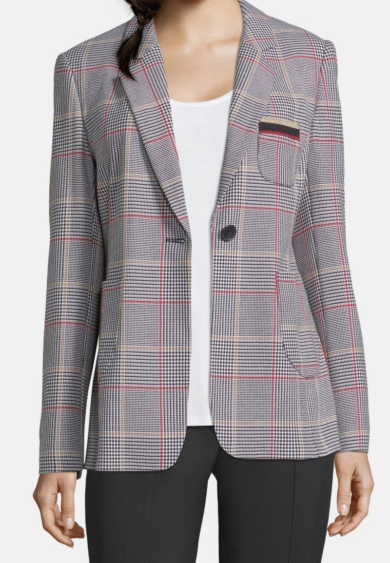 Femme portant un blazer gris à carreaux avec des rayures rouges et beiges sur un haut blanc et un pantalon noir, montrée des épaules aux hanches.