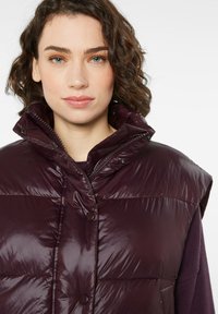 Dunkel-lila Puffer-Weste mit hohem Kragen, glänzender Textur, Druckknopfverschluss und tailliertem Schnitt, ohne Ärmel und mit glatter Oberfläche.