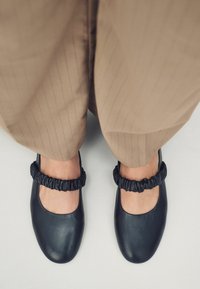 Zwarte leren Mary Jane-schoenen met een ronde neus, voorzien van een gerimpelde zwarte elastische enkelband. Gecombineerd met lichtbruine streepjesbroek.