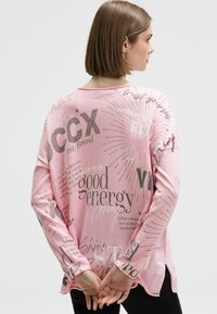 Frau mit kurzen braunen Haaren, die ein lockeres rosa Langarmshirt trägt, bedruckt mit verschiedenen motivierenden Sprüchen, und die die Hände hinter dem Rücken hält.