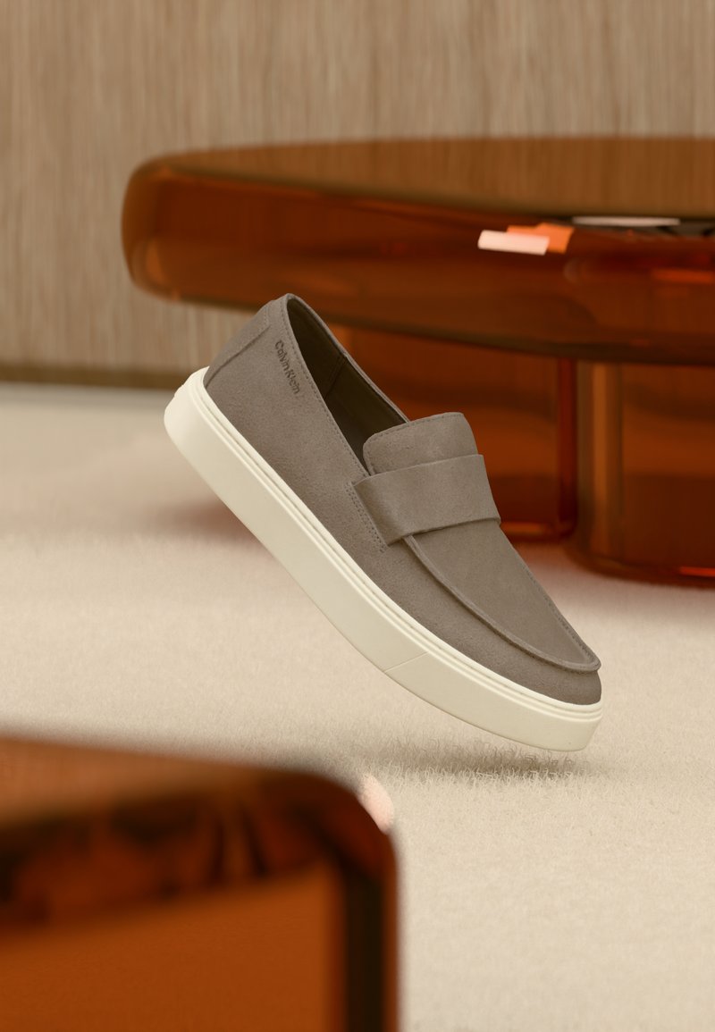 Γκρι σουέτ παπούτσι τύπου slip-on με λευκή σόλα, αιωρούμενο πάνω από μπεζ χαλί, με διαφανή πορτοκαλί έπιπλα στο φόντο.