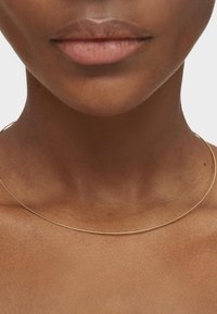 Złoty naszyjnik choker o cienkim, gładkim wzorze spoczywający na obojczyku, podkreślający teksturę skóry i naturalne odcienie.