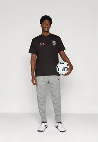 Μαύρο μπλουζάκι με λογότυπο και κόκκινο κείμενο, γκρι αθλητική φόρμα και λευκά αθλητικά παπούτσια. Κρατάει μια μπλε και λευκή ποδοσφαιρική μπάλα.