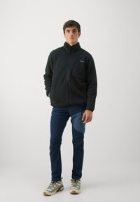 Giacca di pile nera con colletto alto, zip frontale e tasca sul petto. Abbinata a pantaloni blu navy con un pannello testurizzato e scarpe chiare.