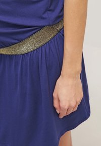 Violetti mekko, jossa on teksturoitu kultavyö, kerätty kangas vyötäröllä, ja käsi lepää hellästi vaatteen päällä. Pehmeä ja sileä materiaali.