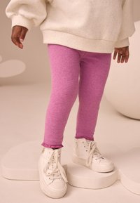 Heledad roosad leggingsid pehme kattega ja volangiga alumisel äärel, valgete kõrgete tossudega hele-taustal.