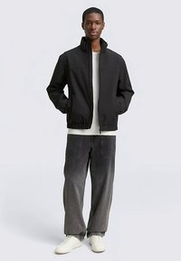 Veste noire à fermeture éclair avec un col haut, présentant un design minimaliste. Associée à un jean gris à jambes larges et des baskets blanches.