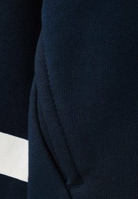 Tkanina mikiny námořnické modré barvy s texturovaným úpletem, doplněná o bílý proužek. Detail ukazuje šití a design bočního švu.