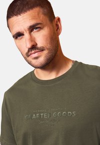 Muž s krátkými vlasy a vousy nosí olivově zelené tričko s textem "Workshop Essentials Crafted Goods Unmatched Comfort" na přední straně.