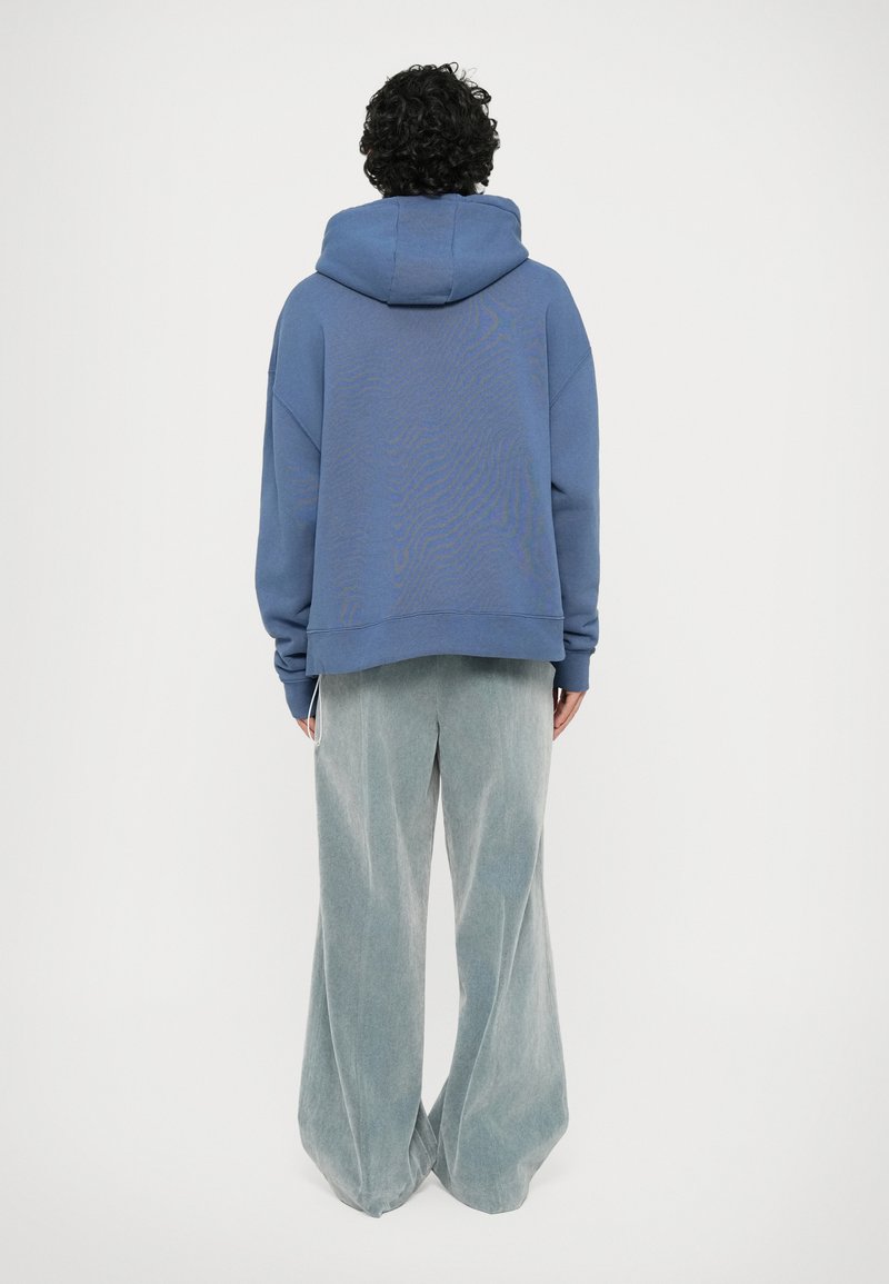 Haut à capuche oversize bleu avec un motif texturé, associé à un pantalon en velours côtelé gris clair à jambes larges, mettant en avant une silhouette décontractée.
