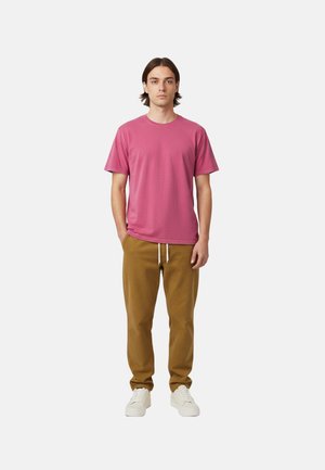 Junger Mann steht mit den Händen in den Taschen, trägt ein rosa T-Shirt, braune Hosen mit Kordelzug und weiße Turnschuhe vor einem einfarbigen Hintergrund.