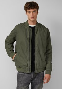 Olivgrüner Bomberjacke mit Frontreißverschluss, gerippten Bündchen und Saum. Über einem schwarzen Pullover getragen, verfügt sie über seitliche Taschen und hat eine glatte Textur.