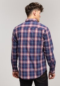 Mężczyzna z krótkimi kręconymi włosami, ubrany w niebieską, czerwoną i białą koszulę w kratę z długim rękawem oraz ciemne dżinsy, pokazany z tyłu.