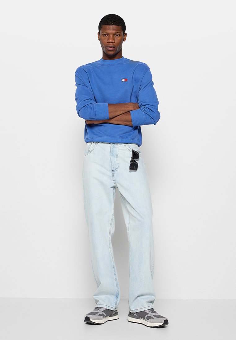 Niebieska bluza z małym logo, jasnoniebieskie dżinsy i szare sneakersy. Stylizacja ma luźny krój i casualowy design.