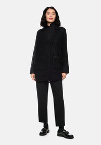 Schwarze perforierte Strickjacke mit Knopfverschluss, zwei Fronttaschen und Reverskragen, kombiniert mit schwarzen Hosen und Schuhen.