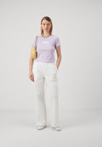 Fialová tričko s krátkym rukávom a bielym logom, kombinované s bielymi cargo nohavicami na elastický pás. Modelka drží malú žltú kabelku.